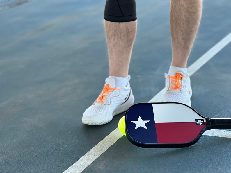 K&ouml;nnte beinhalten: Eine Person in wei&szlig;en Sneakers mit orangenen Schn&uuml;rsenkeln steht auf einem Pickleball-Platz mit einem Pickleball-Schl&auml;ger, der das Design der Flagge des Bundesstaates Texas tr&auml;gt. Ein gelber Pickleball liegt auf dem Platz.