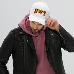 May include: White baseball cap with the letters "SWT" in maroon and gold. The cap has a curved brim and a structured fit. The hat is worn by a person wearing a black leather jacket and a pink hoodie.
