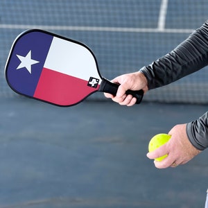 K&ouml;nnte beinhalten: Eine Person h&auml;lt einen gelben Pickleball und einen blau-rot-wei&szlig;en Pickleball-Schl&auml;ger mit dem Design der Texas-Flagge. Der Schl&auml;ger hat einen schwarzen Griff.