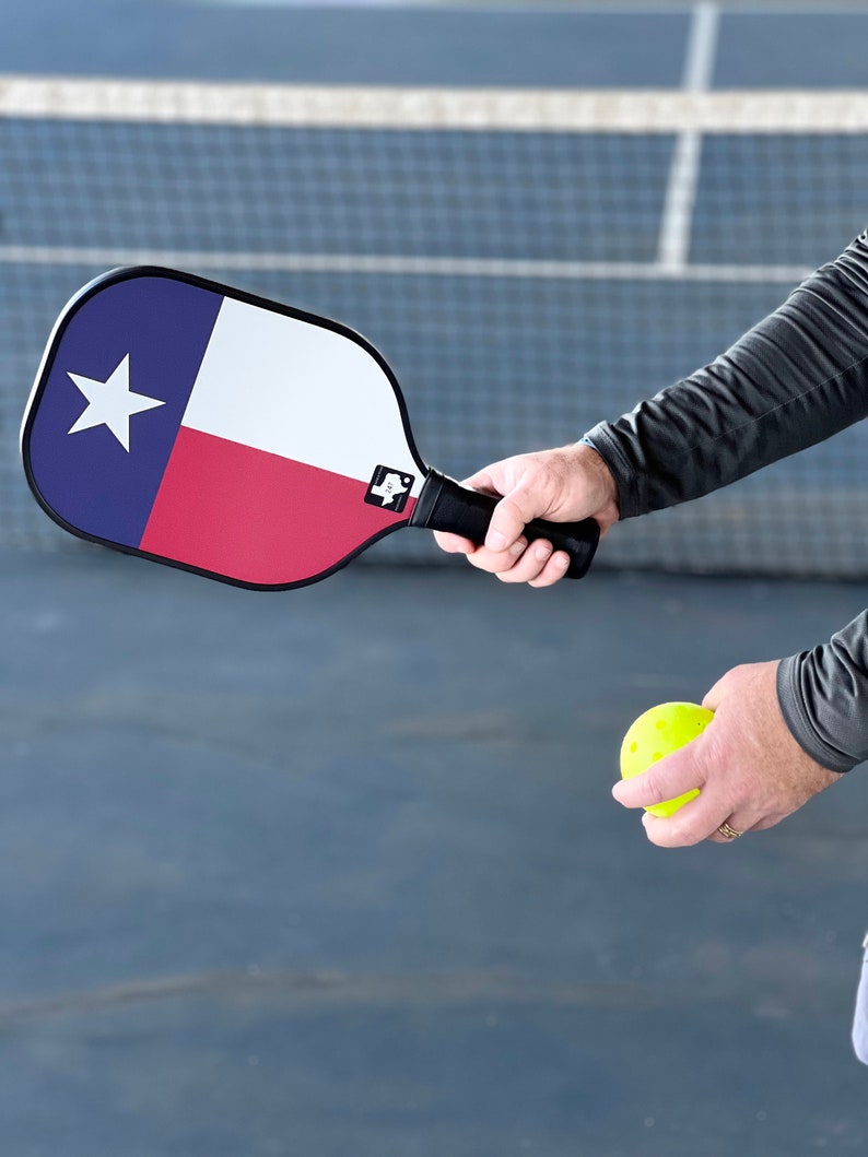 K&ouml;nnte beinhalten: Eine Person h&auml;lt einen gelben Pickleball und einen blau-rot-wei&szlig;en Pickleball-Schl&auml;ger mit dem Design der Texas-Flagge. Der Schl&auml;ger hat einen schwarzen Griff.