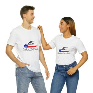 May include: Two white t-shirts with a graphic of a boat on Lake LBJ, Texas. The boat has a Texas flag on the back. The shirts are worn by two people.