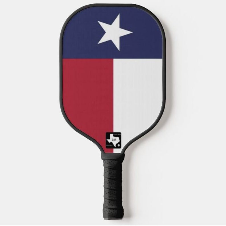 K&ouml;nnte beinhalten: Ein schwarz-wei&szlig;er Pickleball-Schl&auml;ger mit einem Texas-Flaggen-Design. Der Schl&auml;ger hat einen schwarzen Griff und eine schwarz-wei&szlig;e Umrandung des Bundesstaates Texas mit der Nummer 247 im Inneren.