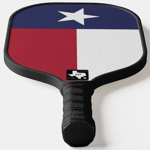 K&ouml;nnte beinhalten: Ein schwarz-wei&szlig;er Pickleball-Schl&auml;ger mit einem Texas-Flaggen-Design. Der Schl&auml;ger hat einen schwarzen Griff mit einer strukturierten Oberfl&auml;che und ein schwarz-wei&szlig;es Logo mit dem Text "247" und einer Silhouette des Bundesstaates Texas.