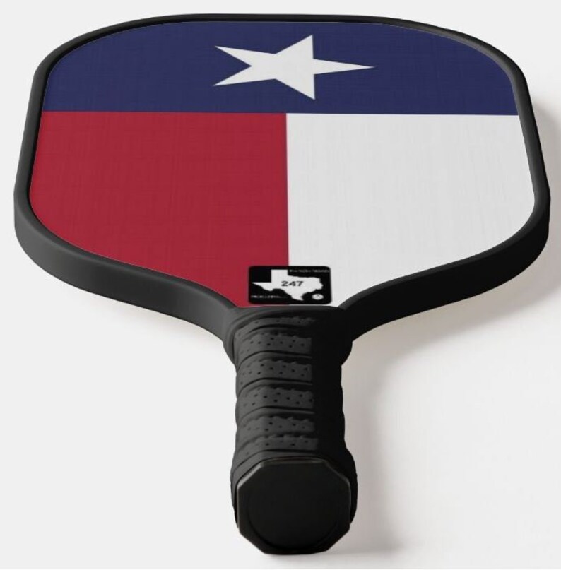 K&ouml;nnte beinhalten: Ein schwarz-wei&szlig;er Pickleball-Schl&auml;ger mit einem Texas-Flaggen-Design. Der Schl&auml;ger hat einen schwarzen Griff mit einer strukturierten Oberfl&auml;che und ein schwarz-wei&szlig;es Logo mit dem Text "247" und einer Silhouette des Bundesstaates Texas.