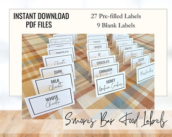 May include: Printable s'mores bar food labels in black and white text on white cardstock. The labels are arranged on a plaid tablecloth. The text on the labels includes "Chocolate", "Dark", "Milk Chocolate", "White Chocolate", "Caramel", "Snicker", "Toasted", "GF", "Cinnamon", "Honey Graham Crackers", "Sliced Strawberries", and "Marshmallows". The text "27 Pre-filled Labels" and "9 Blank Labels" is visible at the top of the image.