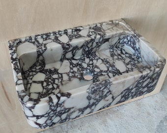 Puede incluir: Un lavabo de mármol blanco con vetas negras. El lavabo es rectangular y tiene un borde redondeado. Está montado en una pared.