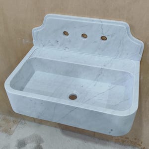 May include: A white marble wall-mounted sink with a rectangular basin and a decorative backsplash. The sink features three circular holes and a central drain. The marble has gray veining throughout, adding a natural pattern to the fixture.
