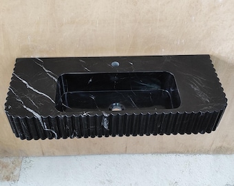 May include: A black marble bathroom sink with a rectangular basin and a scalloped edge design. The sink has a single faucet hole and a drain. White veining adds contrast to the dark stone.