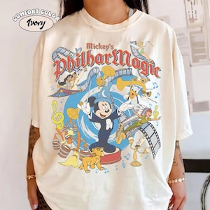 May include: An ivory-coloured short-sleeve t-shirt featuring a graphic print titled Mickey's PhilharMagic. The design includes Mickey Mouse in a blue wizard hat, Simba, Tinker Bell, and Peter Pan surrounded by musical notes and film strips.