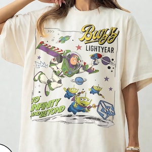 Puede incluir: Una camiseta de color marfil con un estampado gráfico de Buzz Lightyear volando con las alas extendidas, acompañado por tres alienígenas verdes. El texto dice Buzz Lightyear y To Infinity and Beyond, rodeado de estrellas, planetas y una superficie lunar.