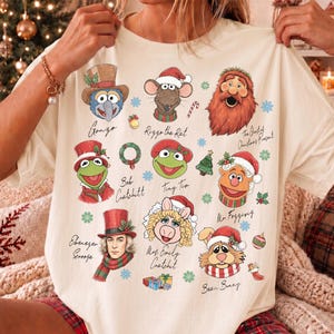 May include: A cream-colored t-shirt featuring a Muppet Christmas Carol graphic with characters including Gonzo, Rizzo the Rat, The Ghost of Christmas Present, Bob Cratchit, Tiny Tim, Mr. Fozzie, Ebenezer Scrooge, Mrs. Emily Cratchit, and Bean Bunny.