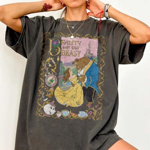Puede incluir: Una camiseta de manga corta gris carbón con un estampado gráfico de La Bella y la Bestia. El diseño incluye a Bella con un vestido amarillo, la Bestia con una chaqueta azul y personajes secundarios, enmarcado por filigrana dorada y el texto Beauty and the Beast.