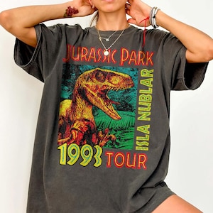 May include: A charcoal gray short-sleeve t-shirt featuring a graphic of a dinosaur in a jungle setting. The text reads JURASSIC PARK, ISLA NUBLAR, 1993, and TOUR in red, yellow, and green lettering. The shirt has a crew neckline and a loose fit.
