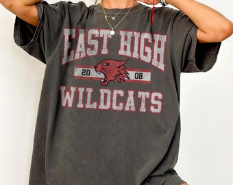 May include: A charcoal grey short-sleeve t-shirt featuring the text EAST HIGH WILDCATS in a distressed red and white collegiate font. A red wildcat mascot graphic is centred between the words with the numbers 20 and 08 on either side.