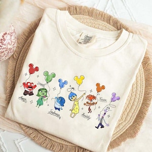 May include: Cream-colored crew neck t-shirt featuring cartoon characters holding Mickey Mouse-shaped balloons. The characters represent emotions like Anger, Disgust, Sadness, Joy, Anxiety, and Fear. The shirt has the Comfort Colors brand tag.