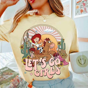 May include: Yellow t-shirt with a graphic of a cowgirl riding a horse, surrounded by cacti, sunflowers, and a cowboy hat. The text "LET'S GO GIRLS" is in pink bubble letters.