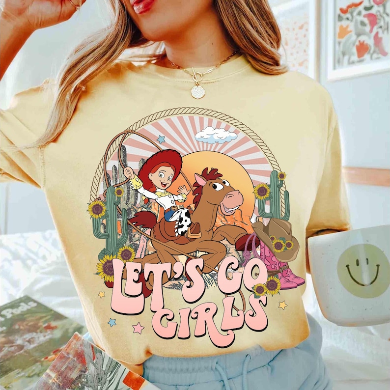 May include: Yellow t-shirt with a graphic of a cowgirl riding a horse, surrounded by cacti, sunflowers, and a cowboy hat. The text "LET'S GO GIRLS" is in pink bubble letters.