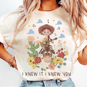 May include: A cream-colored t-shirt featuring a graphic of a cowgirl playing a guitar while riding a horse, surrounded by cacti, flowers, clouds, and musical notes. The text "I KNEW IT I KNEW YOU" is printed in brown letters at the bottom of the design.