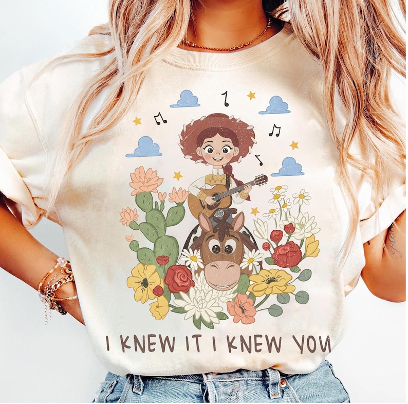 May include: A cream-colored t-shirt featuring a graphic of a cowgirl playing a guitar while riding a horse, surrounded by cacti, flowers, clouds, and musical notes. The text "I KNEW IT I KNEW YOU" is printed in brown letters at the bottom of the design.