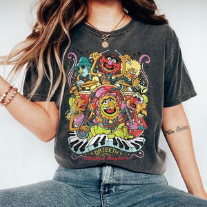 May include: Dark gray t-shirt featuring a colorful graphic of Dr. Teeth and the Electric Mayhem band. The design includes various Muppet characters playing instruments, with the band's name and logo at the bottom. The shirt has a vintage, worn look.