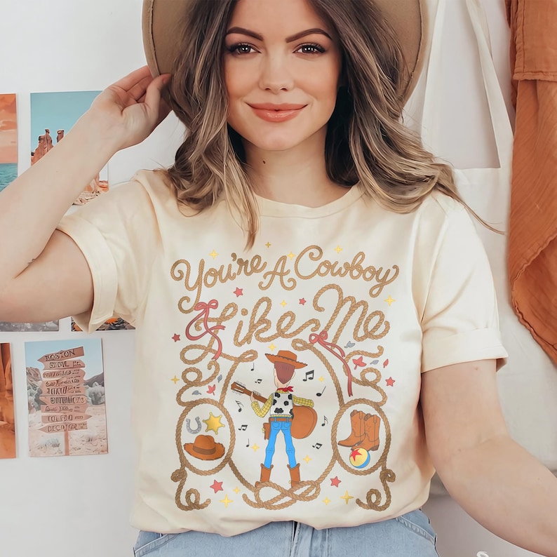 May include: A cream-colored short-sleeve t-shirt featuring a graphic print with the text "You're A Cowboy Like Me" in brown script. The design includes a central cowboy figure playing a guitar, surrounded by a lasso, boots, and stars.