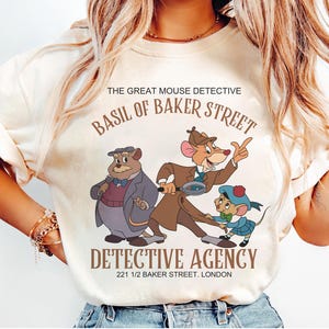 May include: A cream-coloured t-shirt featuring a graphic of characters from The Great Mouse Detective. The text reads THE GREAT MOUSE DETECTIVE, BASIL OF BAKER STREET, DETECTIVE AGENCY, and 221 1/2 BAKER STREET. LONDON. The design includes three mice.