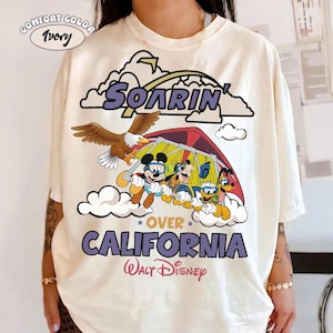 May include: An ivory t-shirt featuring a graphic print of Mickey Mouse, Goofy, Donald Duck, and Pluto hang gliding. The design includes a bald eagle, clouds, and the text Soarin' Over California and Walt Disney. A Comfort Color Ivory label is visible.