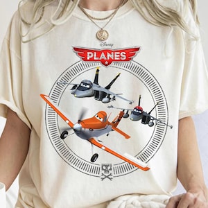 May include: Cream-colored t-shirt featuring the Disney Planes logo and illustrations of Dusty Crophopper and other aircraft. The design includes a circular graphic with the word "PLANES" in red and white.