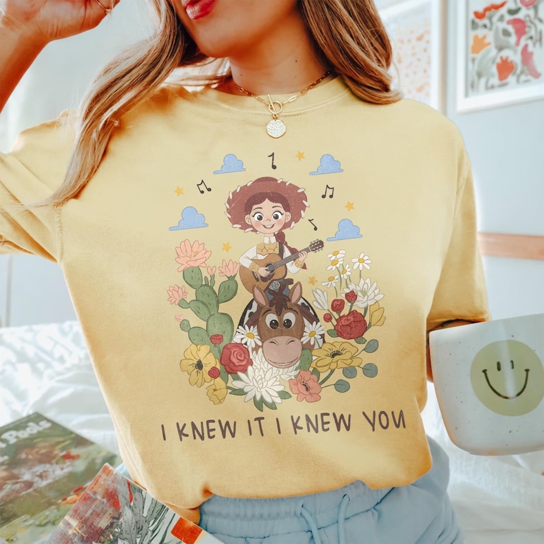 May include: A pale yellow t-shirt featuring a graphic print of a cartoon cowgirl playing a guitar while riding a horse, surrounded by cacti, flowers, clouds, and musical notes. The text "I KNEW IT I KNEW YOU" is printed in dark brown below the graphic.