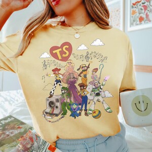 May include: A pale yellow t-shirt featuring a graphic print of various animated characters, including a cowgirl, a space ranger, and a dinosaur. Above the characters is a red heart with the letters TS and musical notes. The design includes clouds.