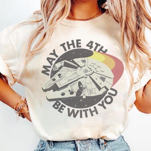 May include: Cream-coloured t-shirt with a graphic of the Millennium Falcon spaceship and the text "MAY THE 4TH BE WITH YOU" in a circular design. The design includes yellow, red, and black accents, giving a vintage aesthetic.