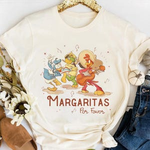 May include: Cream-colored t-shirt featuring a cartoon band of three characters, including Donald Duck, playing instruments. The text "Margaritas Por Favor" is printed in brown script below the characters. The shirt is styled with blue jeans and fall decor.