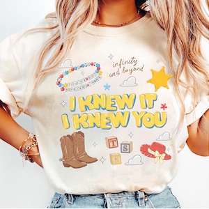 May include: A cream-colored t-shirt featuring a graphic print with the text I KNEW IT I KNEW YOU, infinity and beyond, and YOU'VE GOT A FRIEND IN ME. The design includes cowboy boots, a sheriff star, alphabet blocks, and a red cowboy hat.