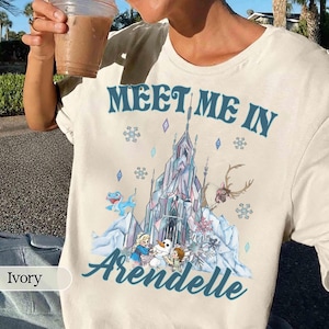 May include: Ivory-colored t-shirt featuring the text "MEET ME IN Arendelle" in blue, with a graphic of a castle, snowflakes, and cartoon characters. The shirt is a crew neck and has short sleeves.