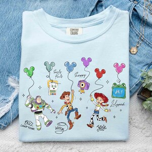 Op de afbeelding: Lichtblauw T-shirt met stripfiguren Buzz Lightyear, Woody en Jessie, met Mickey Mouse-vormige ballonnen. Het shirt heeft de tekst "Buzz Lightyear", "Woody", "Jessie", "Smarty Pants", "Happy" en "Lilypad".