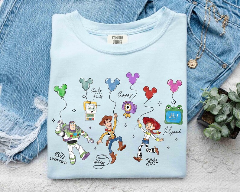 Op de afbeelding: Lichtblauw T-shirt met stripfiguren Buzz Lightyear, Woody en Jessie, met Mickey Mouse-vormige ballonnen. Het shirt heeft de tekst "Buzz Lightyear", "Woody", "Jessie", "Smarty Pants", "Happy" en "Lilypad".