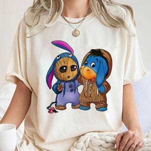 May include: Cream-colored t-shirt featuring two cartoon characters dressed in costumes. One is a wooden-like figure in a blue bunny costume, and the other is a blue donkey in a brown hooded outfit. The characters are hugging each other.