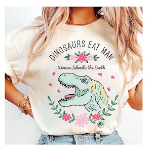 Pode incluir: Camiseta creme com o texto "DINOSAURS EAT MAN" acima de uma ilustração de dinossauro de desenho animado. O texto "Woman Inherits the Earth" está abaixo do texto e do dinossauro. O dinossauro é cercado por flores.
