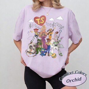 May include: A light purple t-shirt featuring a graphic print of various animated characters, a red heart with the letters TS, musical notes, and a vintage camera. The design includes a colourful array of figures in diverse outfits, clouds, and a ball.