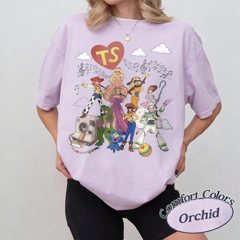 May include: A light purple t-shirt featuring a graphic print of various animated characters, a red heart with the letters TS, musical notes, and a vintage camera. The design includes a colourful array of figures in diverse outfits, clouds, and a ball.