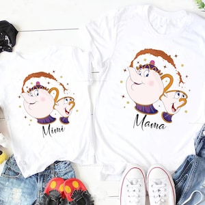 May include: White t-shirts with a cartoon teapot and teacup design, and the words "Mimi" and "Mama" in black script. The design includes gold stars and brown accents. The shirts are shown with denim shorts, shoes, and soft toys.