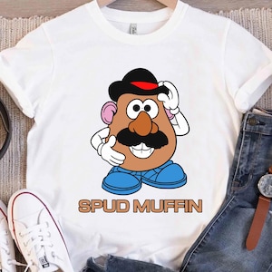 Könnte beinhalten: Weißes T-Shirt mit einem Cartoon-Mr. Potato Head-Design, einschließlich schwarzem Hut, Schnurrbart und blauen Schuhen. Der Schriftzug "SPUD MUFFIN" ist in Braun aufgedruckt. Das T-Shirt hängt an einem Kleiderbügel.