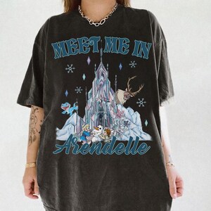 May include: Dark gray t-shirt featuring a graphic from the movie Frozen. The design includes the text "Meet Me In Arendelle" above an illustration of the castle, characters, and snowflakes. The shirt is a short-sleeved crew neck.