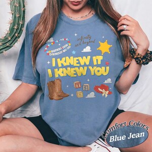 May include: A blue denim-colored t-shirt featuring a graphic print with the text I KNEW IT I KNEW YOU, infinity and beyond, and a beaded bracelet. The design includes illustrations of brown cowboy boots, alphabet blocks, a yellow star, and a red hat.