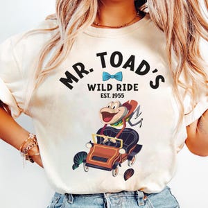 Könnte beinhalten: Weißes T-Shirt mit einem Cartoon-Bild von Mr. Toad, der ein Auto fährt. Der Text auf dem Shirt lautet "Mr. Toad's Wild Ride Est. 1955".