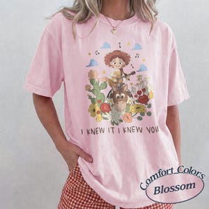 May include: A light pink t-shirt featuring a graphic of a cartoon cowgirl playing a guitar while riding a horse, surrounded by cacti and flowers. The text "I KNEW IT I KNEW YOU" is printed below the illustration. The shirt has short sleeves.