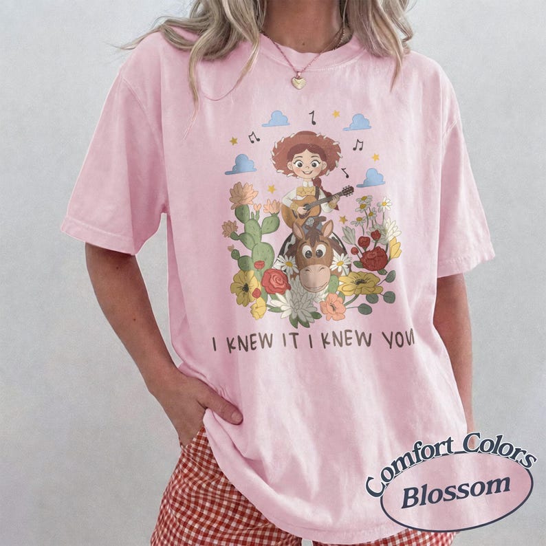 May include: A light pink t-shirt featuring a graphic of a cartoon cowgirl playing a guitar while riding a horse, surrounded by cacti and flowers. The text "I KNEW IT I KNEW YOU" is printed below the illustration. The shirt has short sleeves.