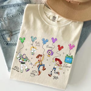 Op de afbeelding: Cr&egrave;mekleurig T-shirt met stripfiguren Buzz Lightyear, Woody en Jessie die Mickey Mouse-vormige ballonnen vasthouden. Op het shirt staan de woorden "Buzz Lightyear", "Woody" en "Jessie". Een bruine hoed en een blauwe spijkerbroek staan op de achtergrond.