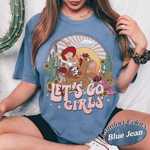 May include: Blue jean-colored t-shirt featuring a cartoon graphic of a cowgirl riding a horse, with the text "LET'S GO GIRLS". The design includes cacti, sunflowers, and a sunset. The shirt is a Comfort Colors brand.