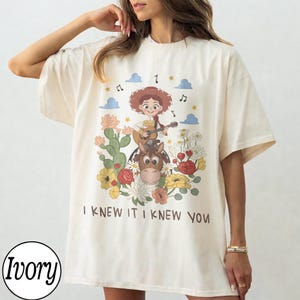 May include: An ivory t-shirt featuring a graphic print of a cartoon character with red hair playing a guitar while riding a brown horse. The design includes colorful flowers, cacti, blue clouds, musical notes, and the text I KNEW IT I KNEW YOU.